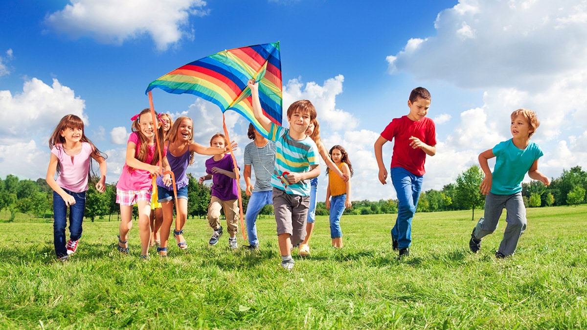 Group of children running across a grassy park beneath a colorful kite.